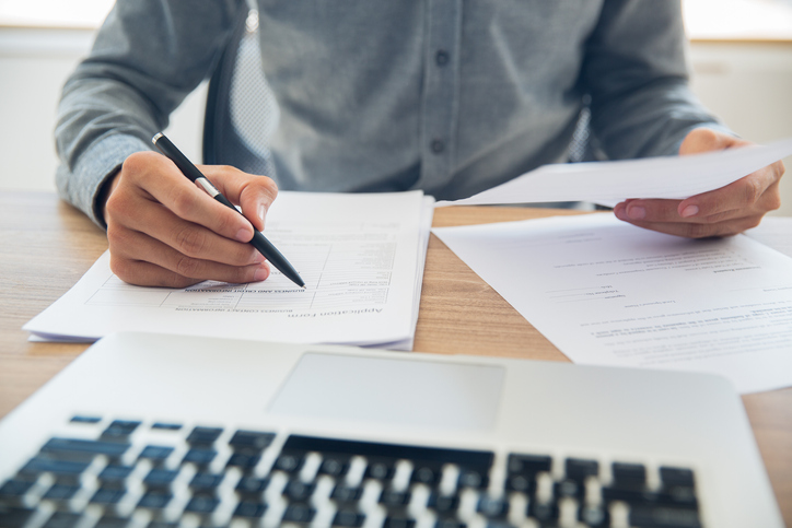 Unrecognizable businessman sitting at table holding ballpoint pen and papers and checking documents in office