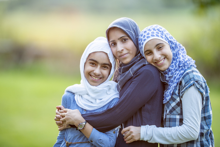 Three Muslim siblings of different ages are outdoors in a park. They are embracing and smiling for the camera.