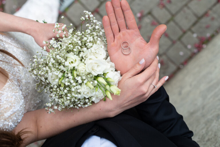 A bride holding a bouquet of white flowers while the groom holds two wedding rings in his palm.