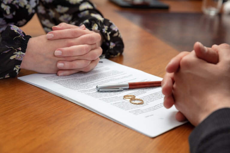 Two people sit across from each other with folded hands over a document, a pen, and two wedding rings on the table, symbolizing divorce proceedings.