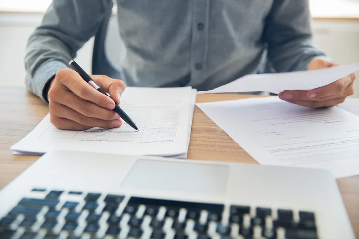 Unrecognizable businessman sitting at table holding ballpoint pen and papers and checking documents in office