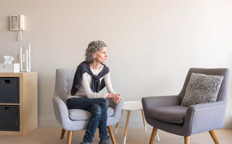 A woman sitting alone on a chair, looking thoughtful and pensive in a quiet room.