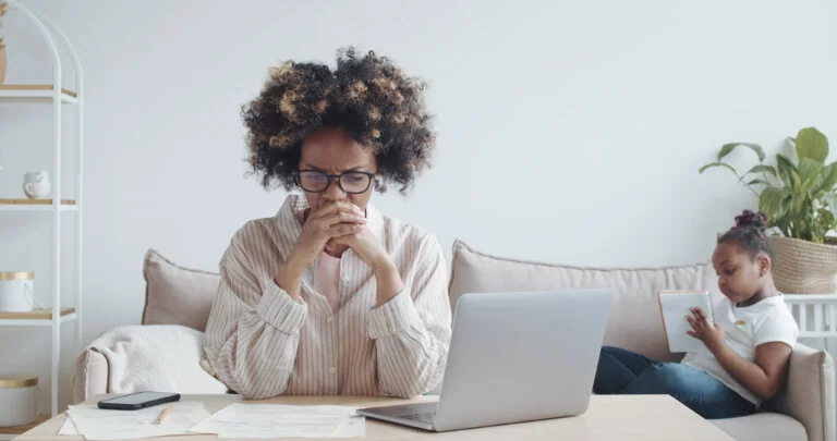 A worried woman sits at a table with papers and a laptop, while a child reads a notebook on the couch in the background.