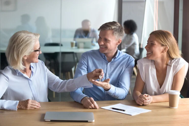 Female real estate agent give keys to excited caucasian couple buying first home together, smiling husband and wife sit at Real Estate Agent office become owners of own apartment. Ownership concept
