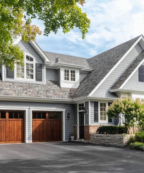 MA USA - A large grey and white modern farmhouse with two wooden garage doors, and beautiful landscaping.